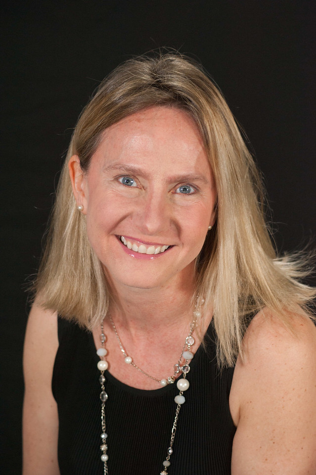 Image of a woman with blonde hair and blue eyes, smiling at the camera. She is wearing a black shirt and silver necklace, and is in front of a black background.