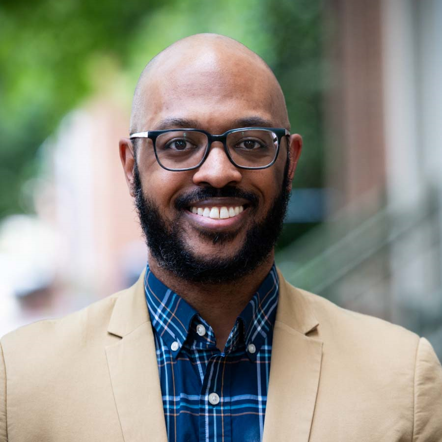 Image of a man smiling at the camera. He is wearing glasses and a blue patterned collared shirt and a tan suit jacket.