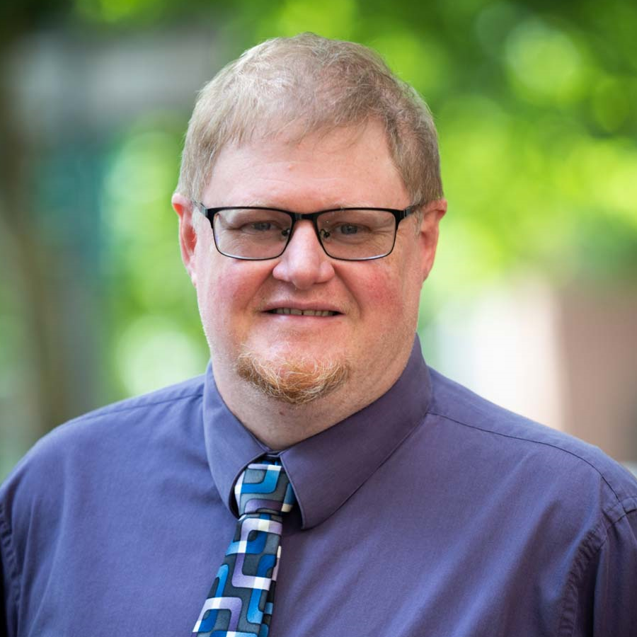 Image of a man with light brown hair who is smiling at the camera. He is wearing glasses and a blue collared shirt, and a blue patterned tie.