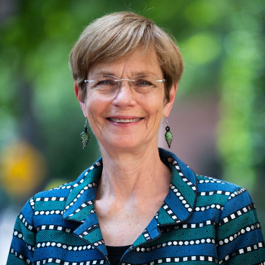 Image of a woman smiling at the camera, she is wearing a teal blue patterned blazer jacket, wearing earrings and glasses.