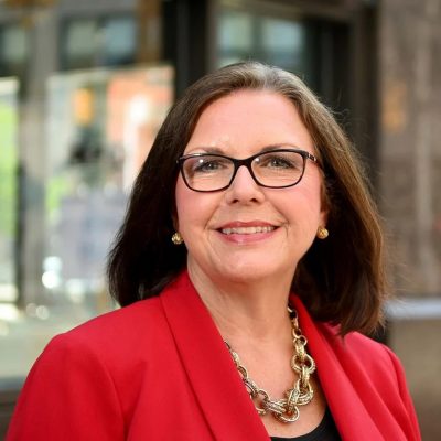 Image of a woman with brown hair, who is wearing glasses and a red blazer, smiling at the camera.