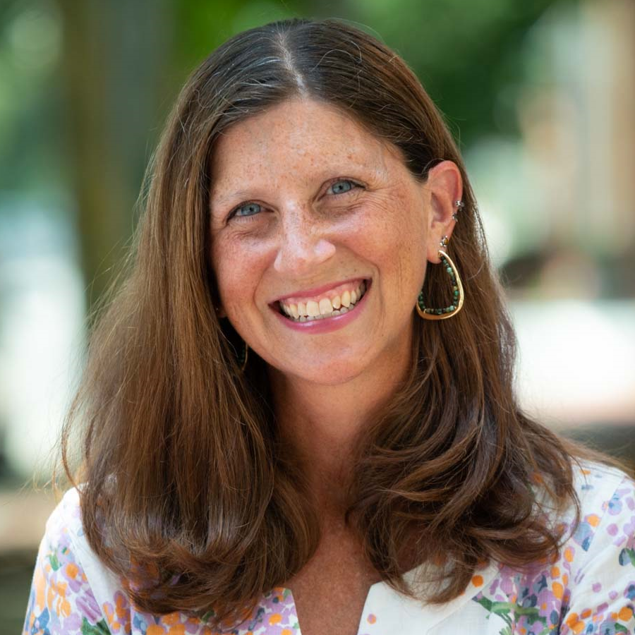 Image of a woman with brown hair smiling at the camera. She is wearing hoop earrings and a white blouse with a colorful pattern.