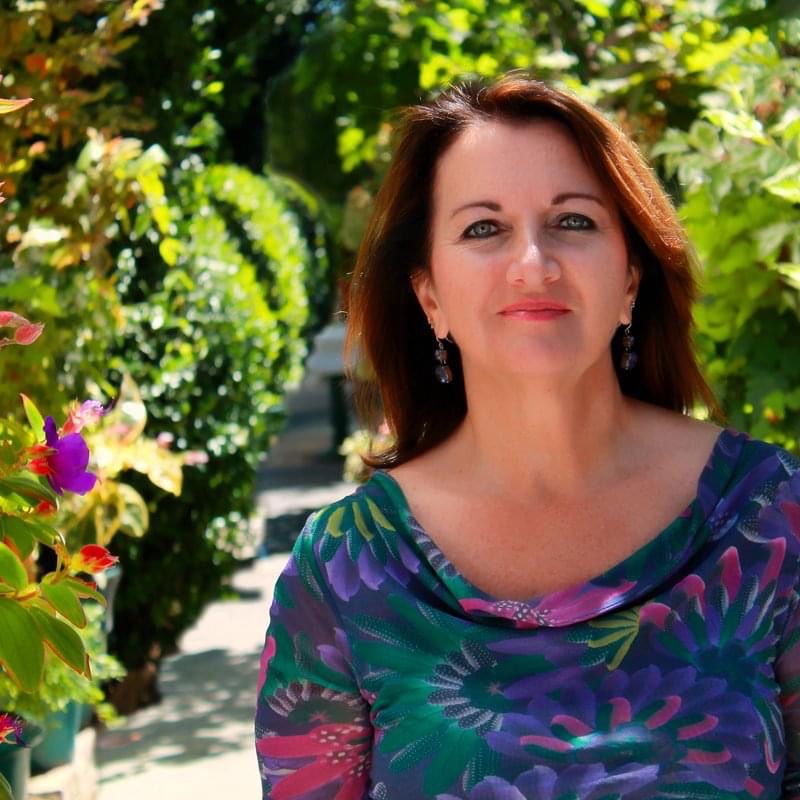 Image of a woman with brown hair wearing a colorful floral blouse, standing in front of greenery and colorful flowers.