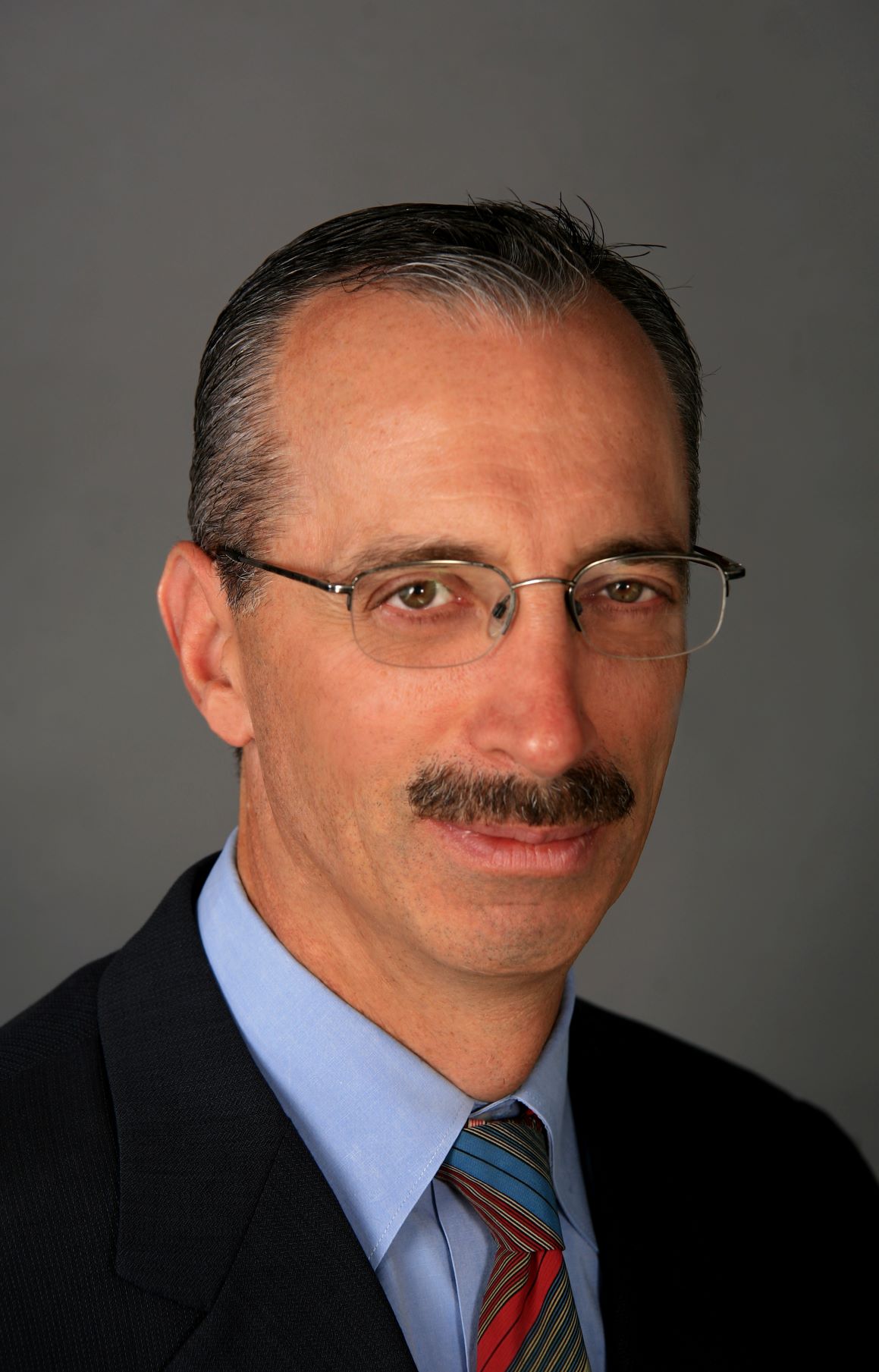 Image of a man with dark hair wearing glasses, a blue collared shirt, red tie and black suit. He is positioned in front of a dark grey background.