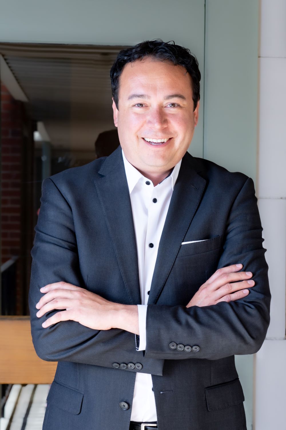 Image of a man with dark hair smiling a the camera, with his arms crossed in front of him. He is wearing a white collared shirt and a black suit jacket.