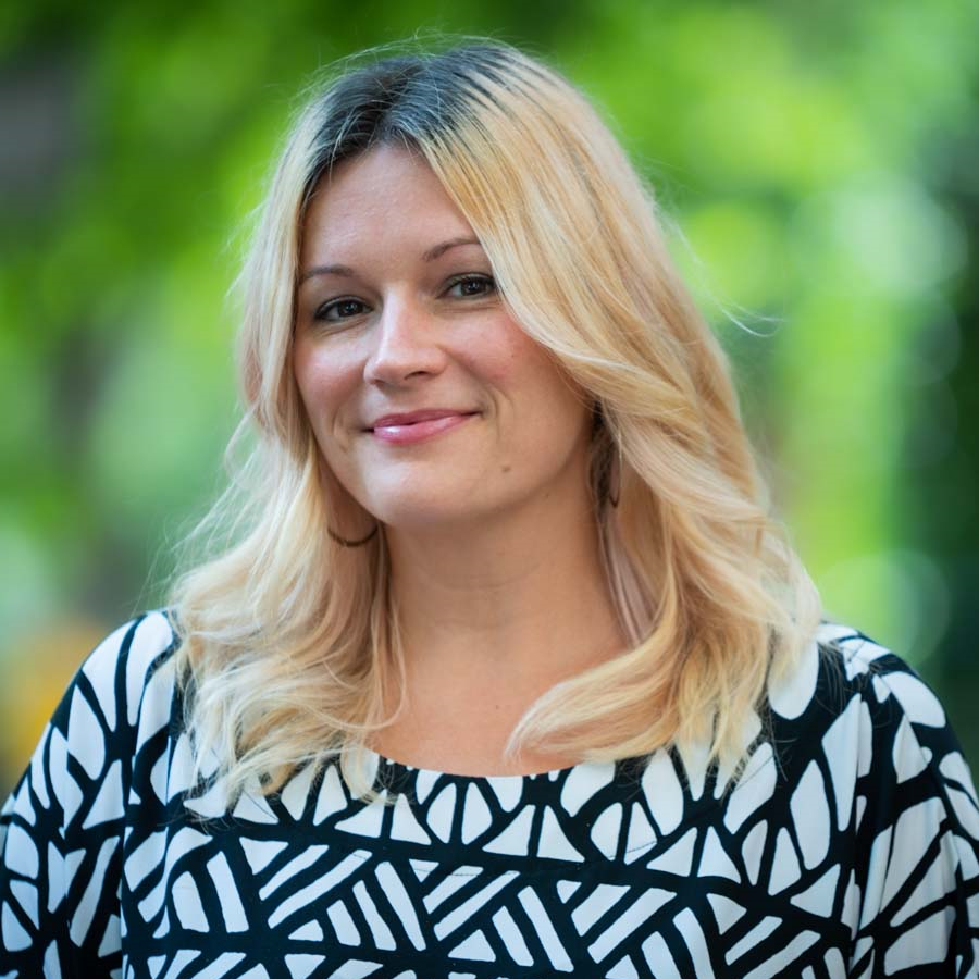 Image of a woman with blonde hair smiling softly at the camera. She is wearing a patterned black and white blouse.