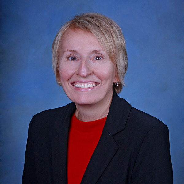 Image of a woman with blonde hair smiling at the camera. She is wearing a red blouse and a black blazer jacket, positioned in front of a blue background.