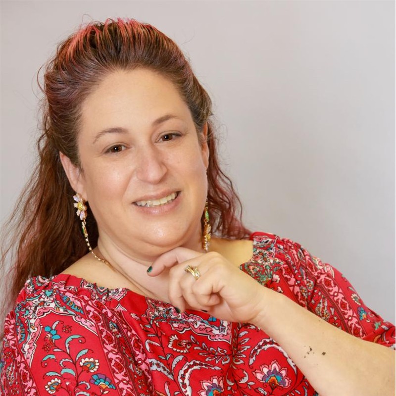 Image of a woman with brown hair smiling at the camera. She is wearing a red patterned blouse and dangling earrings.