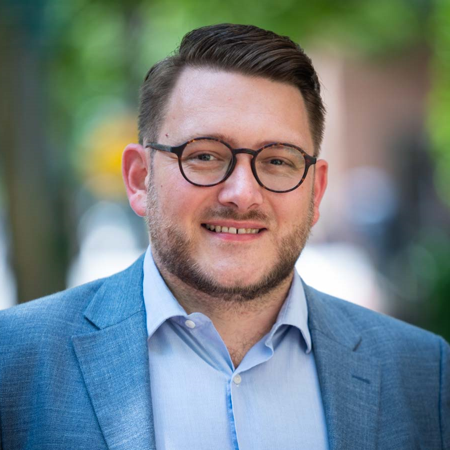 Image of a man with brown hair smiling at the camera, wearing glasses and a light blue collared shirt, and a blue suit jacket.