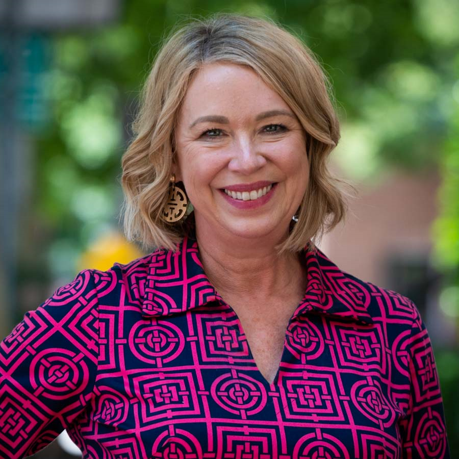 Image of a woman with blonde hair smiling at the camera. She is wearing a patterned pink and navy blue blouse and earrings.