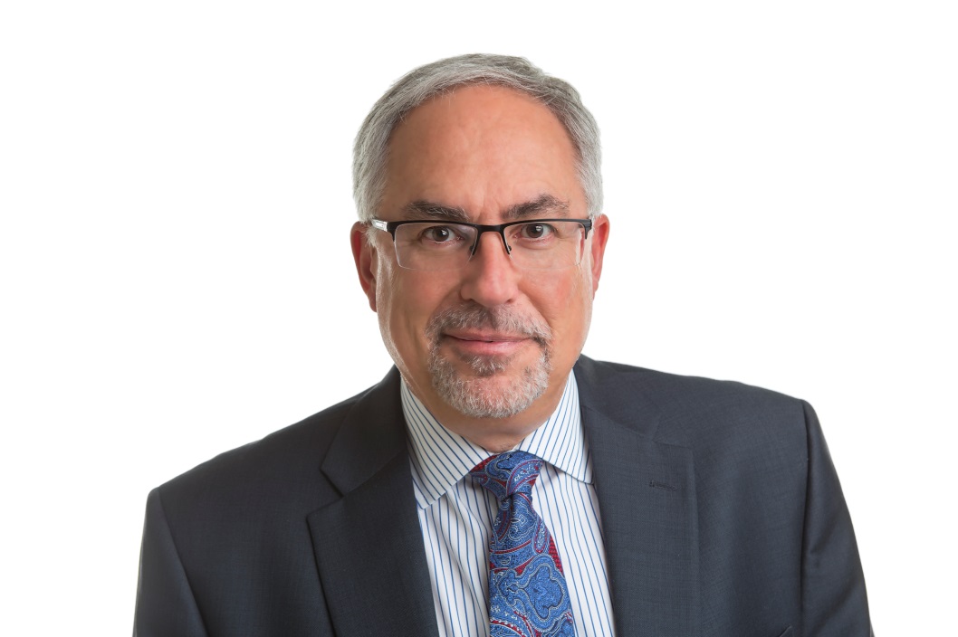 Image of a man wearing glasses smiling softly at the camera. He is wearing a blue tie, collared shirt and suit jacket and is positioned in front of a white background.