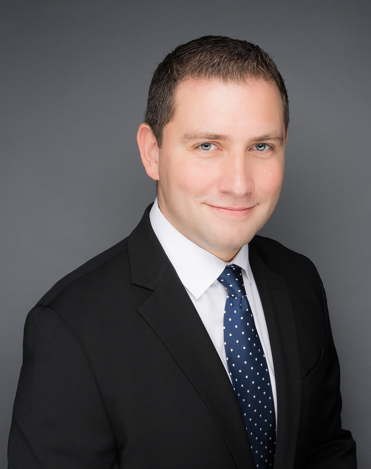 Image of a man with brown hair smiling softly at the camera. He is wearing a white collared shirt, a blue tie and a black suit jacket in front of a dark gray background.