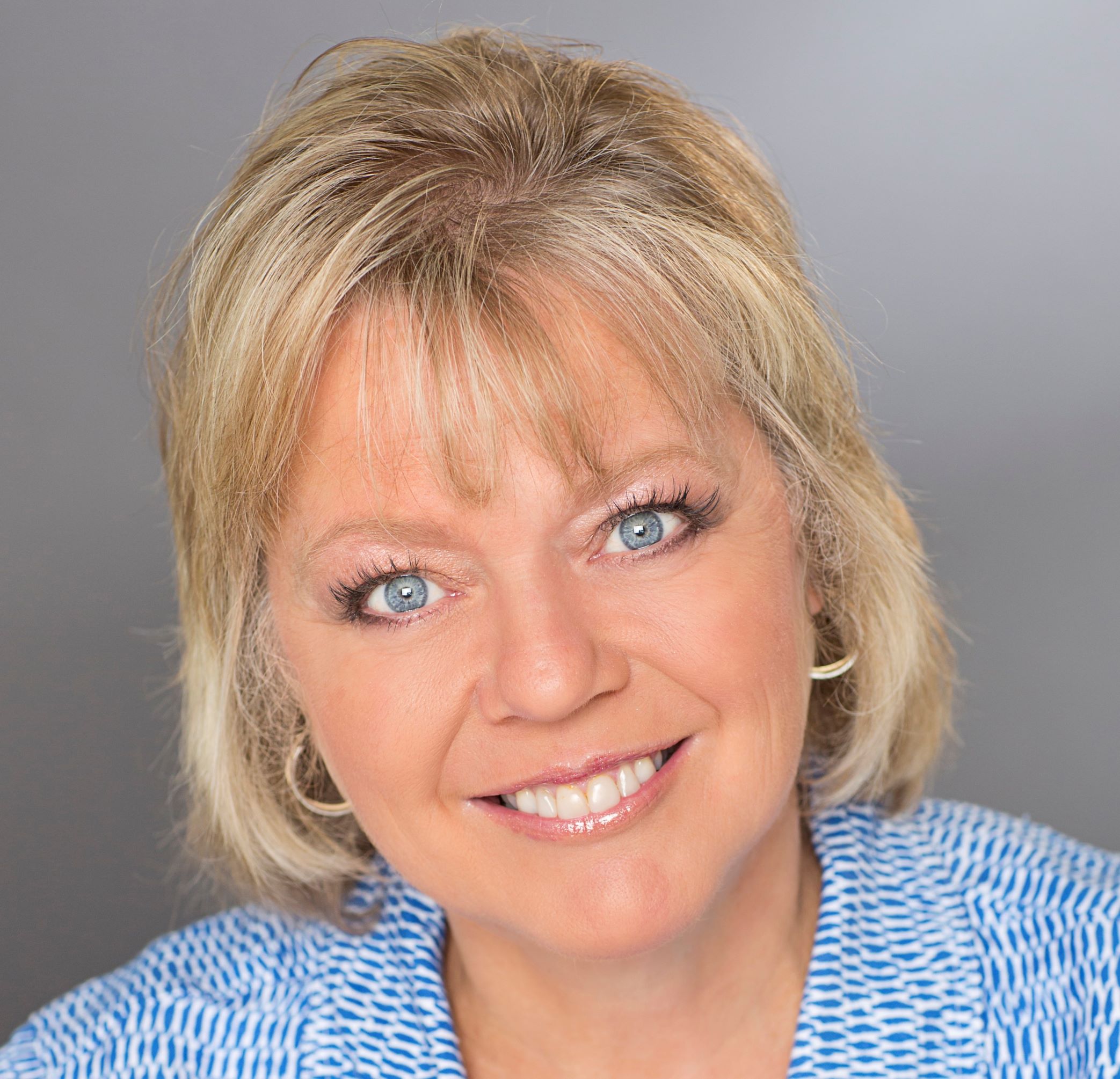 Image of a woman with blonde hair and blue eyes, wearing a blue and white shirt, smiling at the camera in front of a gray background.