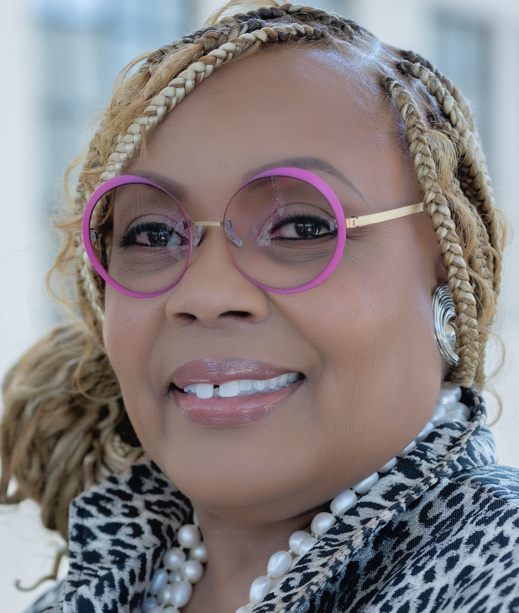 Image of a woman with blonde braids smiling at the camera. She is wearing pink glasses and a leopard print suit jacket.