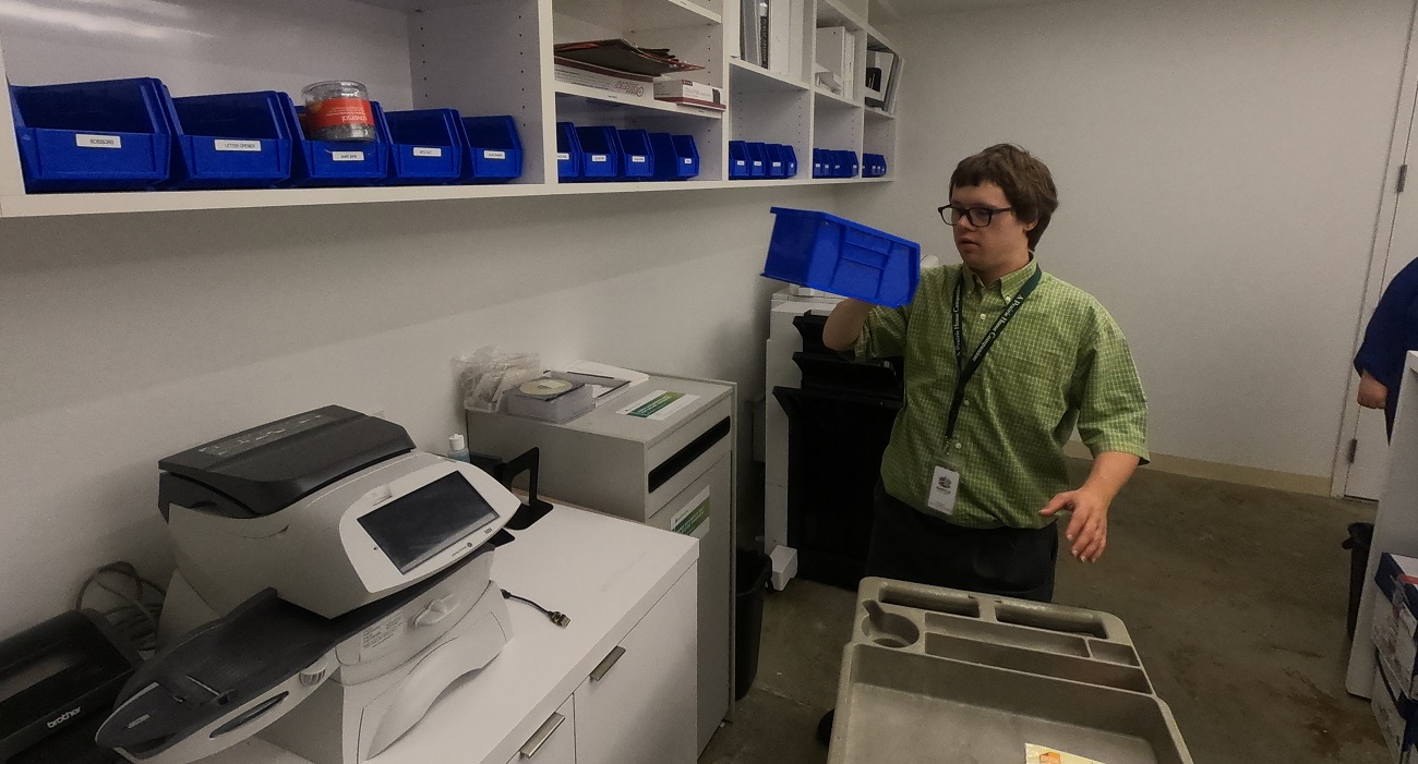 A young man wearing a green shirt and a work badge stocking supplies in the mail room of an office