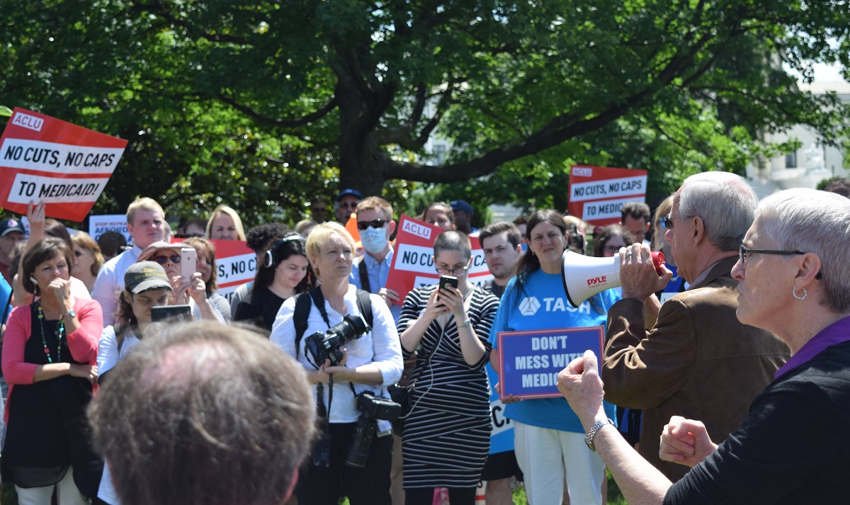 A crowd of supporters at a Medicaid rally holding signs and listening to a Senator speaking through a megaphone.