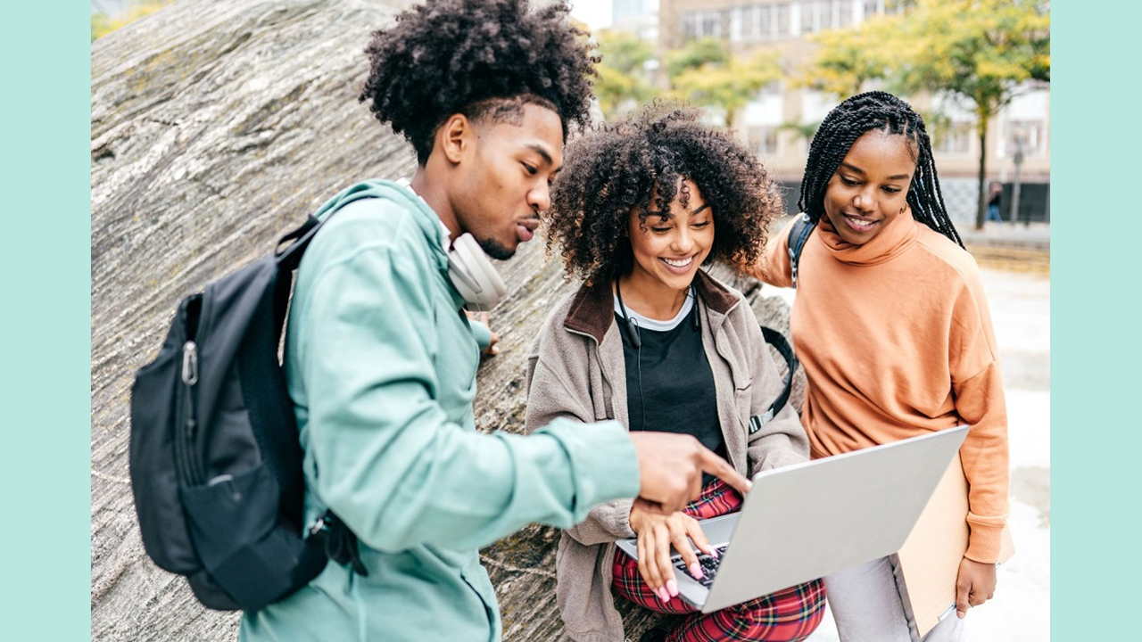 Three students, a man and two women, on a college campus huddling over the laptop of the student in the middle