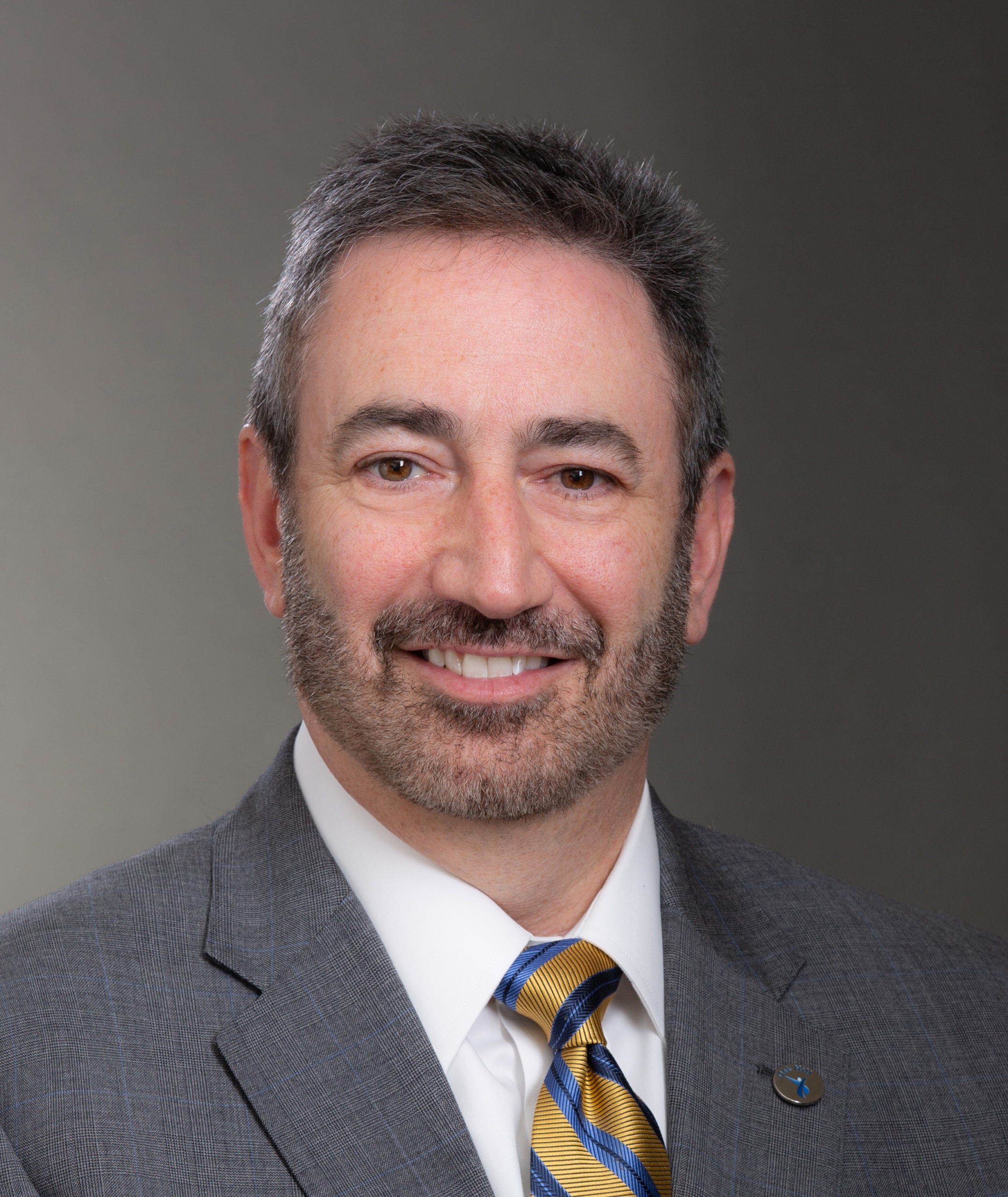 Headshot for Barry Simon, who has short, dark hair and a short beard, and who is wearing a charcoal suit with a white shirt and a striped tie.