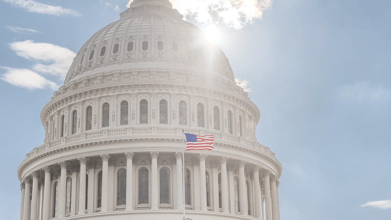 Photo of the top of the U.S. Capitol with the sun shining behind it and an American flag on a flagpole in front
