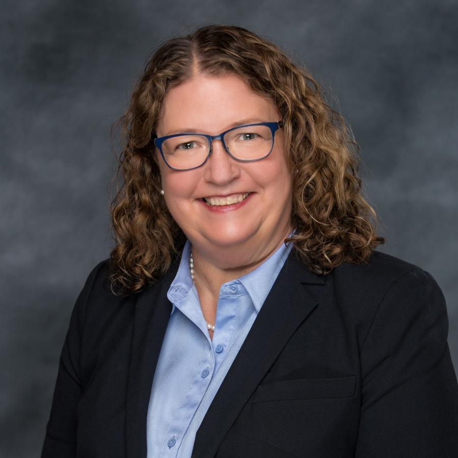 A headshot for Linda Timmons, who has shoulder-length curly hair and is wearing glasses, a blue button-down shirt and a black blazer