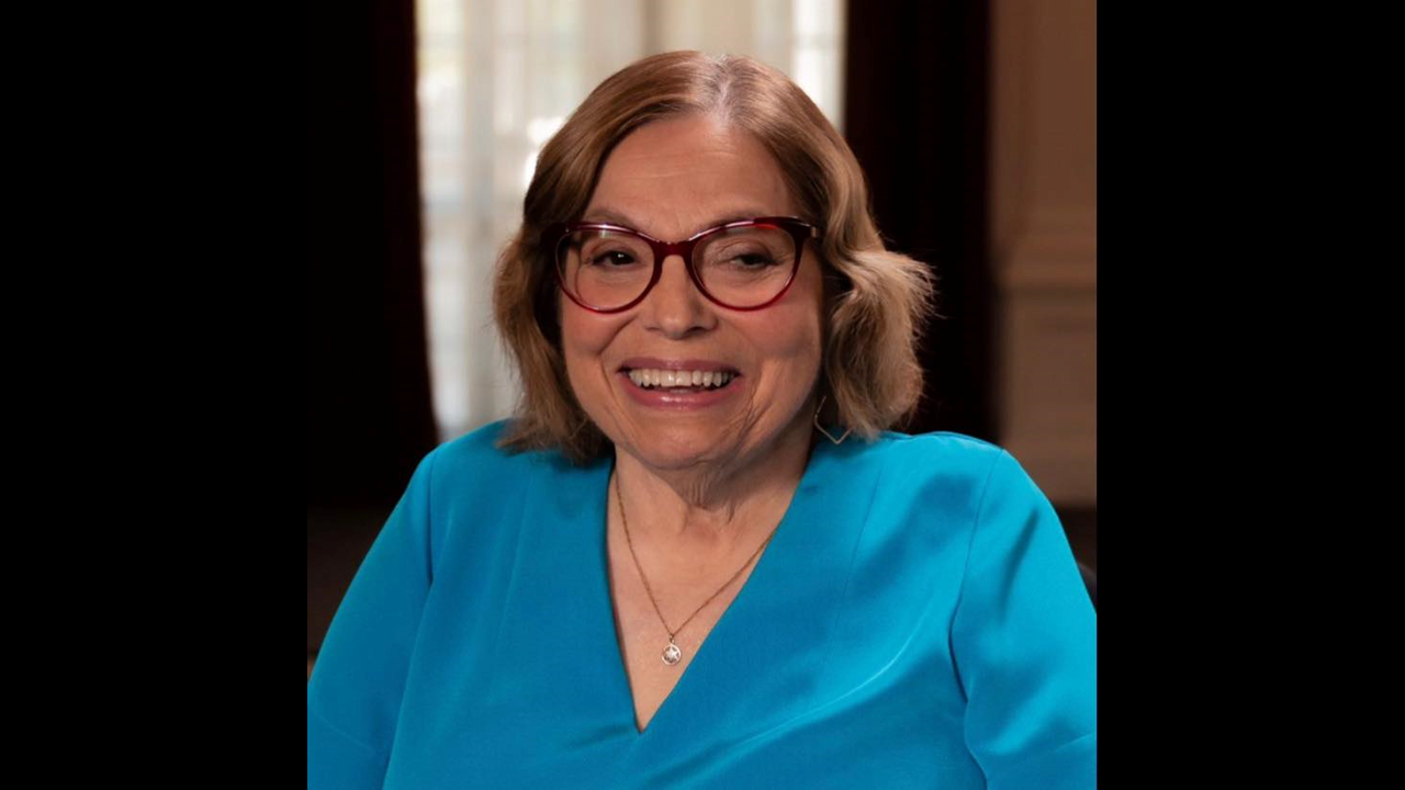 A photo of the late Judy Heumann, who has short brown hair and is wearing maroon-rimmed glasses and a cerulean top.