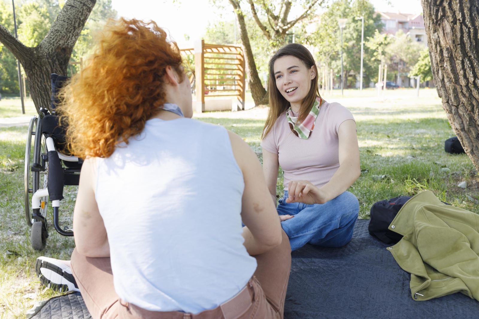 Two women sitting in the park on a blanket facing each other; one of the women is sitting next to her wheelchair.