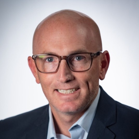 Head shot of Chris Barnes, who is wearing glasses, a blue collared shirt and a dark blazer.