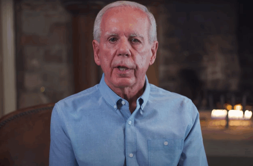 A still image of Tony Coelho wearing a blue shirt and sitting in front of his fireplace speaking to the camera