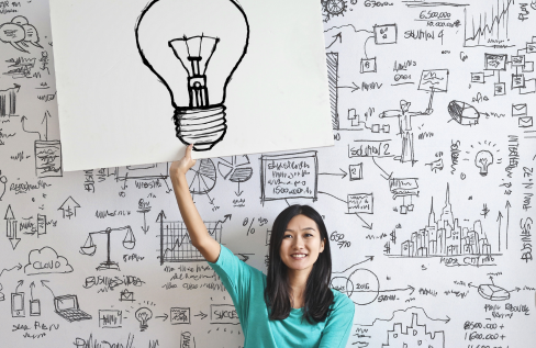 Woman in front of a whiteboard full of drawings holding up a sign with a drawing of a light bulb