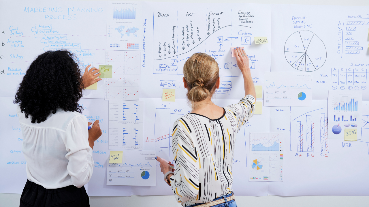 Stock photo of two women in business attire with their backs toward the camera as they point and write on a busy whiteboard