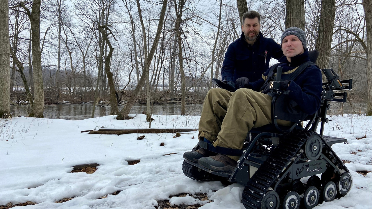 Two brothers posing along a snowy trail, one standing, another in a motorized chair with tracks