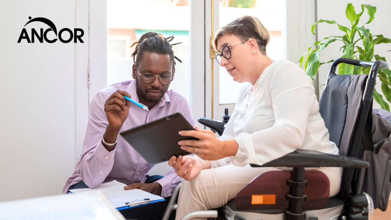 two colleagues look at a tablet together. one is a power wheelchair user