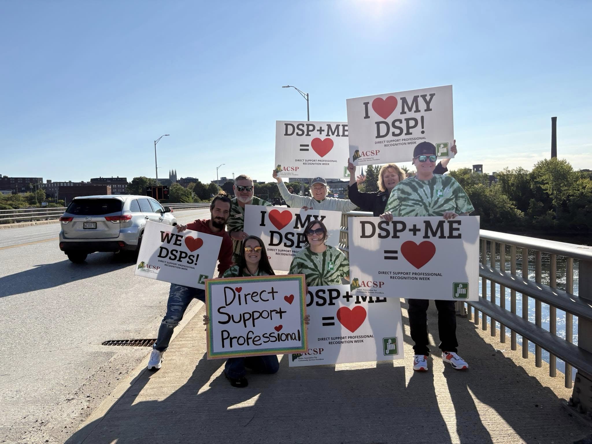 Staff and coworkers stand outside near the road holding big signs that read we heart DSPs