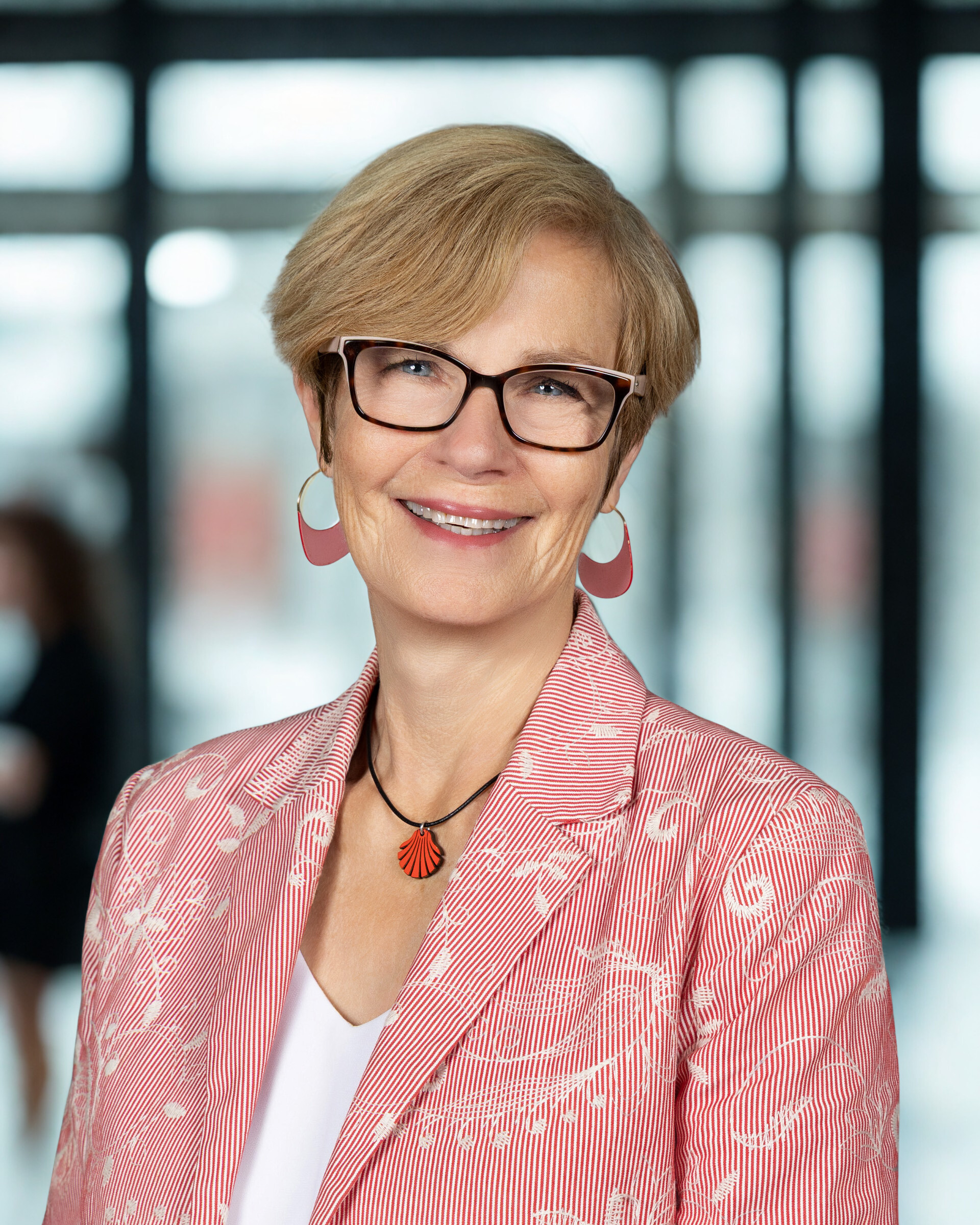 A woman with short blonde hair, black-rimmed glasses, and a red and white blazer smiling for the camera.