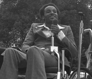 Black and white photo of disability rights activist Brad Lomax speaking at a rally in 1977. Lomax is smiling wearing a plaid suit, sitting in a wheelchair, and holding a microphone.
