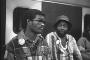 Black and white photo of activists Dennis Billups (left) and Ron Washington (right) during the Section 504 protests in San Francisco. Billups is seated, wearing a plaid jacket, collared shirt, and sunglasses.