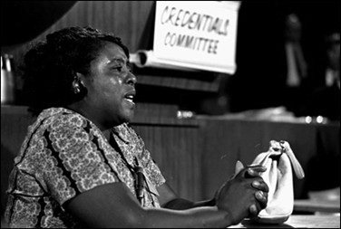 Black and white photo of Fannie Lou Hamer testifying at the 1964 Democratic National Convention in Atlantic City, N.J. Hamer is seated at a table facing the audience with a sign behind her that says “Credentials Committee.”