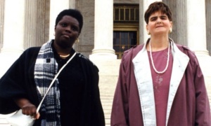 Lois Curtis (left) and Elaine Wilson (right), lead plaintiffs in Olmstead v. L.C., standing on the steps of a courthouse. Curtis is wearing a black coat and plaid scarf and looking at the camera.
