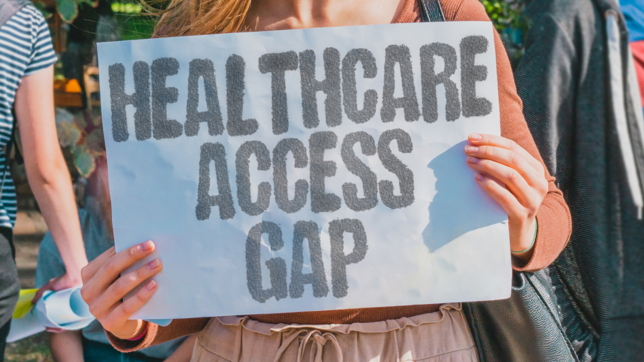 photo of a woman holding a sign at torso height that reads 'HEALTHCARE ACCESS GAP'