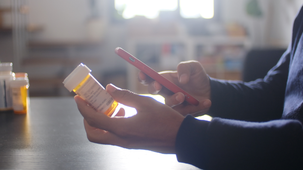 photo of a person holding a prescription bottle in one hand and a smartphone in the other