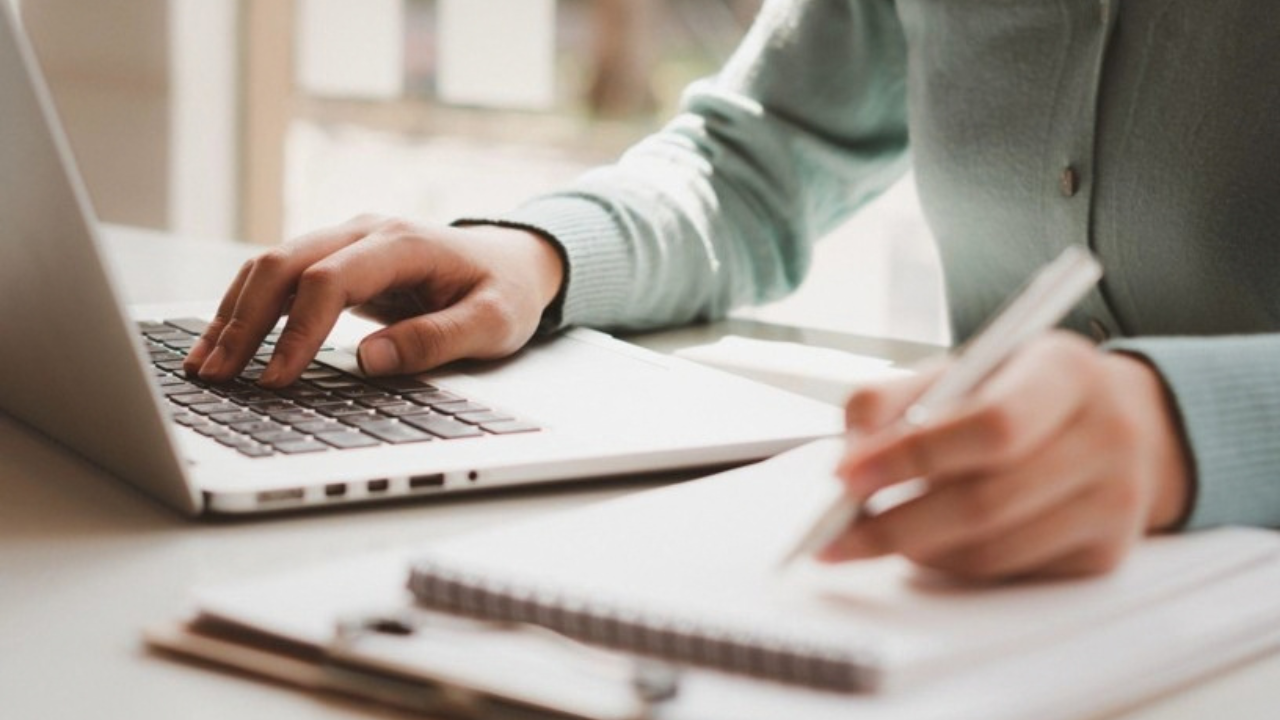 stock photo of one hand typing on a laptop while the other writes with a pen